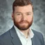 Kevin with short brown hair and a beard is wearing a gray suit jacket and a white polka-dot shirt, embodying the confidence of transactional advisory expertise, posing against a gray textured background.