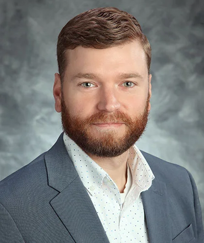 Kevin with short brown hair and a beard is wearing a gray suit jacket and a white polka-dot shirt, embodying the confidence of transactional advisory expertise, posing against a gray textured background.