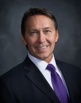 Kevin Bearley, a man in a dark suit with a white shirt and purple tie, smiles confidently against a dark gray background.