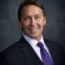 Kevin Bearley, a man in a dark suit with a white shirt and purple tie, smiles confidently against a dark gray background.