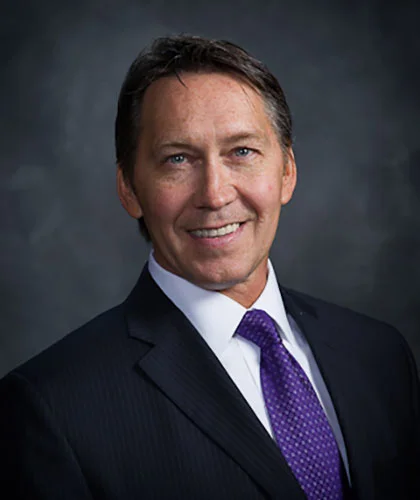 Kevin Bearley, a man in a dark suit with a white shirt and purple tie, smiles confidently against a dark gray background.