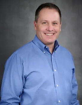 A smiling Mike in a blue button-up shirt, exuding confidence, stands against a gray background, ready to offer expert transactional advisory services.