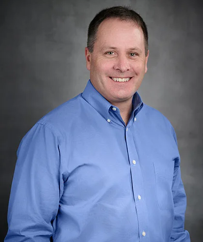 A smiling Mike in a blue button-up shirt, exuding confidence, stands against a gray background, ready to offer expert transactional advisory services.