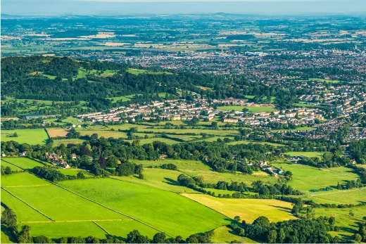 Aerial view of countryside and urban landscape with green fields, scattered trees, and residential areas under a clear blue sky.