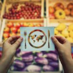 A person holds a card showing a plate with a world map, fork, and knife, with assorted fruits and vegetables blurred in the background.