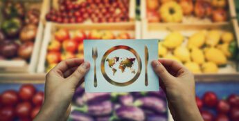 A person holds a card showing a plate with a world map, fork, and knife, with assorted fruits and vegetables blurred in the background.