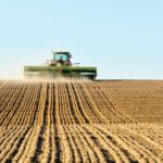 A tractor plows a large, brown field under a clear blue sky, leaving evenly spaced rows of tilled soil behind it.