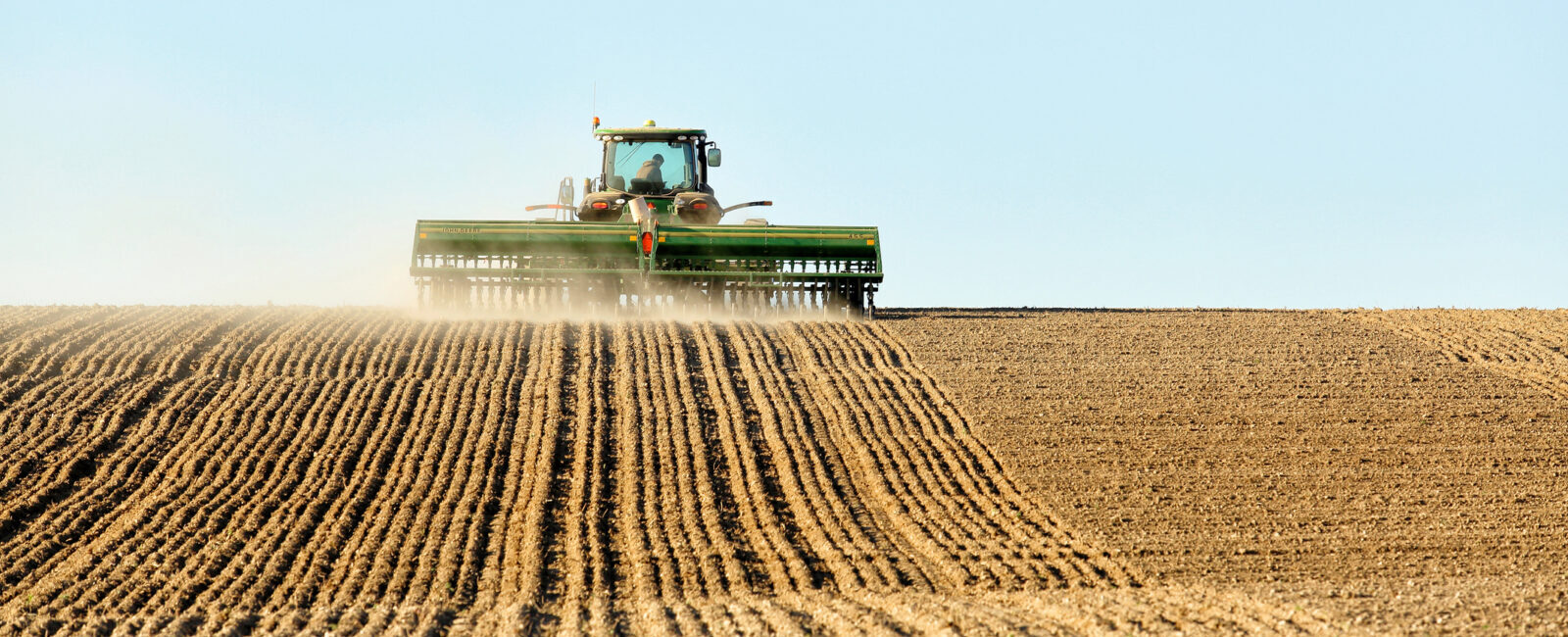 A tractor plows a large, brown field under a clear blue sky, leaving evenly spaced rows of tilled soil behind it.