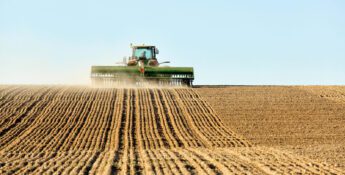 A tractor plows a large, brown field under a clear blue sky, leaving evenly spaced rows of tilled soil behind it.
