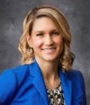 Jessica Dwyer, a woman with shoulder-length blonde hair, smiles while wearing a blue blazer and patterned blouse against a gray background.