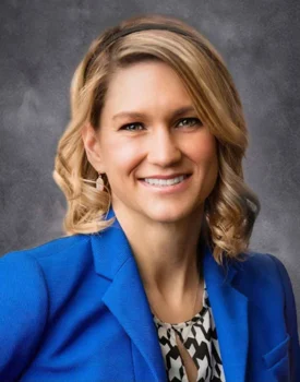 Jessica Dwyer, a woman with shoulder-length blonde hair, smiles while wearing a blue blazer and patterned blouse against a gray background.
