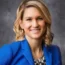 Jessica Dwyer, a woman with shoulder-length blonde hair, smiles while wearing a blue blazer and patterned blouse against a gray background.