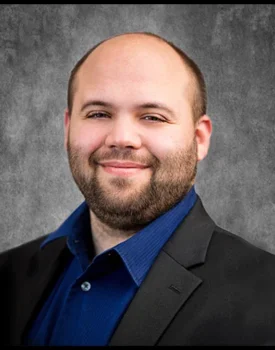 Joseph Brunner, sporting a beard and shaved head, is captured in a dark suit and blue shirt. He offers a slight smile against a gray textured background, exuding confidence and style.