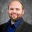 Joseph Brunner, sporting a beard and shaved head, is captured in a dark suit and blue shirt. He offers a slight smile against a gray textured background, exuding confidence and style.