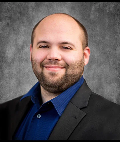 Joseph Brunner, sporting a beard and shaved head, is captured in a dark suit and blue shirt. He offers a slight smile against a gray textured background, exuding confidence and style.