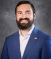 Ryan with a beard smiling, wearing a blue suit with a pin of the U.S. flag lapel, reminiscent of the style favored by accomplished figures like Melissa DeDonder.