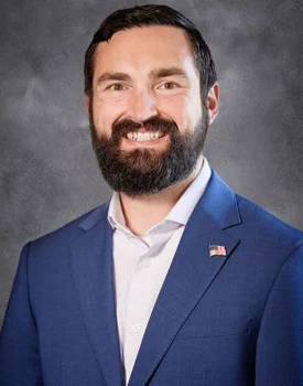 Ryan with a beard smiling, wearing a blue suit with a pin of the U.S. flag lapel, reminiscent of the style favored by accomplished figures like Melissa DeDonder.