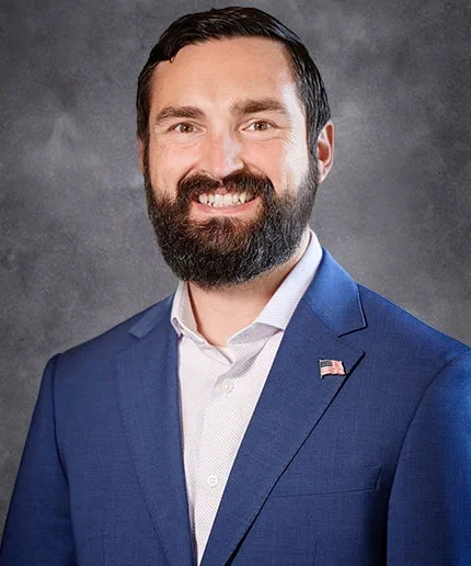 Ryan with a beard smiling, wearing a blue suit with a pin of the U.S. flag lapel, reminiscent of the style favored by accomplished figures like Melissa DeDonder.