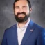 Ryan with a beard smiling, wearing a blue suit with a pin of the U.S. flag lapel, reminiscent of the style favored by accomplished figures like Melissa DeDonder.