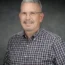 Steve Pitman, a man with short gray hair, glasses, and a goatee, smiles warmly while wearing a checkered shirt against a gray background.
