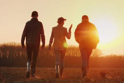 Three people in boots and hats walk through a field at sunrise.