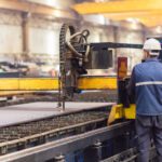 A worker wearing a safety helmet operates a CNC plasma cutting machine in an industrial factory setting.