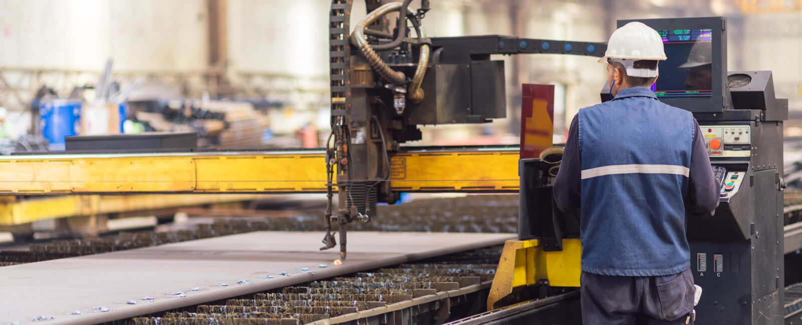 A worker wearing a safety helmet operates a CNC plasma cutting machine in an industrial factory setting.