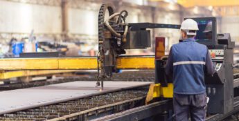 A worker wearing a safety helmet operates a CNC plasma cutting machine in an industrial factory setting.