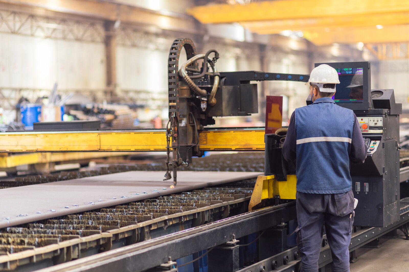 A worker wearing a safety helmet operates a CNC plasma cutting machine in an industrial factory setting.