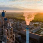 An industrial facility with smoke emitting from a tall chimney, surrounded by fields and a small town under a setting sun.
