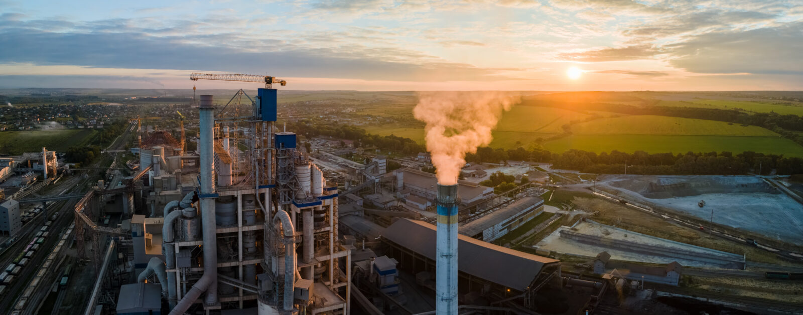 An industrial facility with smoke emitting from a tall chimney, surrounded by fields and a small town under a setting sun.