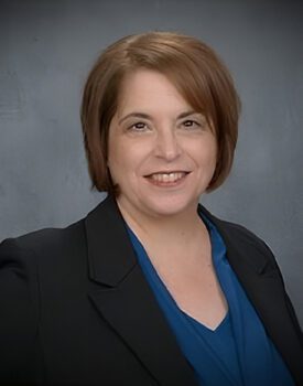 Debra Agnew, a woman with short brown hair wearing a dark blazer and blue blouse, smiles warmly in front of a plain gray background.