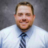 A man with short brown hair and a beard, wearing a light blue dress shirt and a striped tie, poses in front of a gray mottled background.