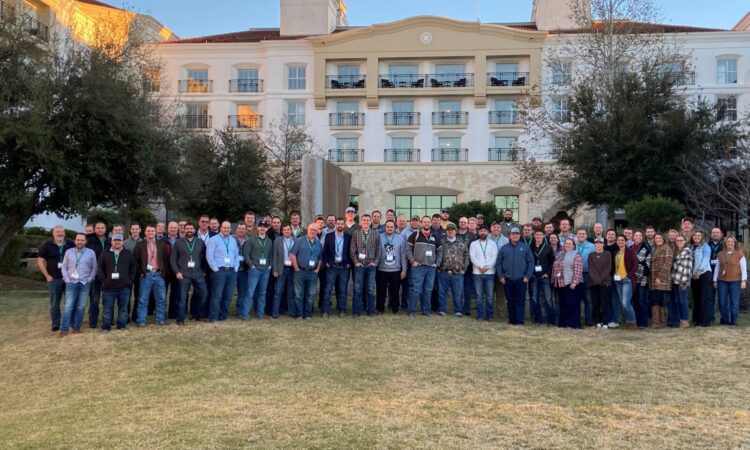 A large group of people posing for a photo on a lawn in front of a multi-story hotel or resort building.