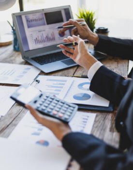 Two people in business attire analyze financial charts and graphs on paper and a laptop, using a calculator and pens at a wooden desk.