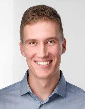 A young man with short light brown hair smiles at the camera, wearing a light blue collared shirt against a plain, light background.