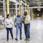 Three people in work attire walk and talk in a spacious industrial facility with large machinery and bright yellow safety posts.