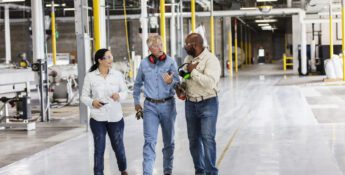 Three people in work attire walk and talk in a spacious industrial facility with large machinery and bright yellow safety posts.