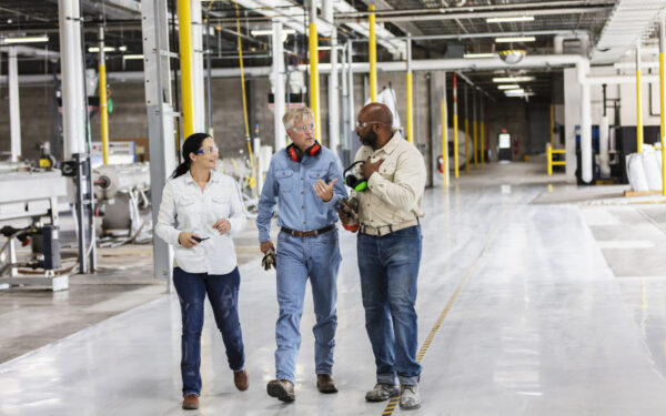 Three people in work attire walk and talk in a spacious industrial facility with large machinery and bright yellow safety posts.
