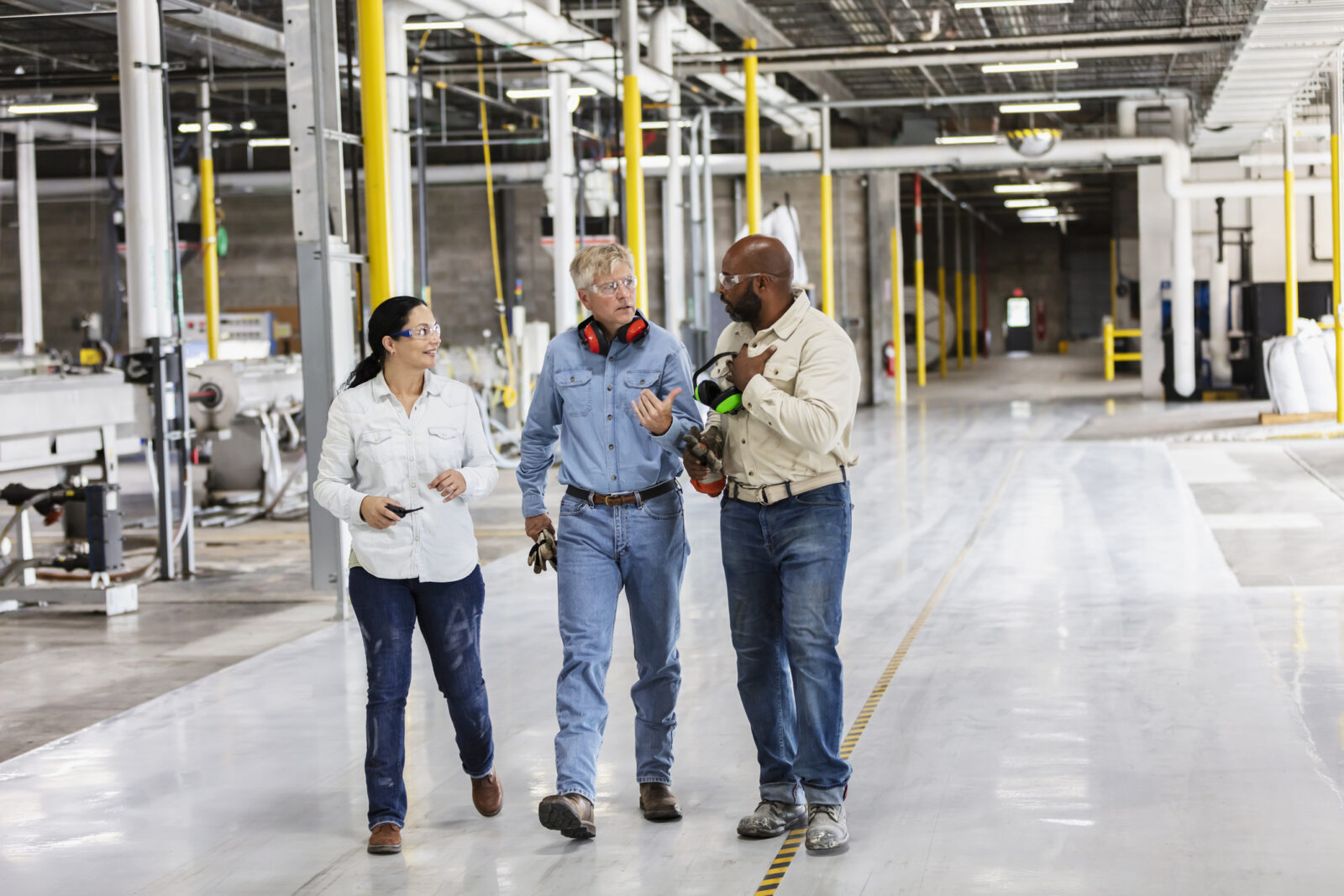 Three people in work attire walk and talk in a spacious industrial facility with large machinery and bright yellow safety posts.