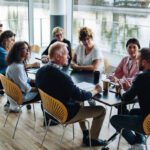 Eight people sit around a table in a modern meeting room with large windows overlooking water, discussing the advantages of peer groups for business leaders.