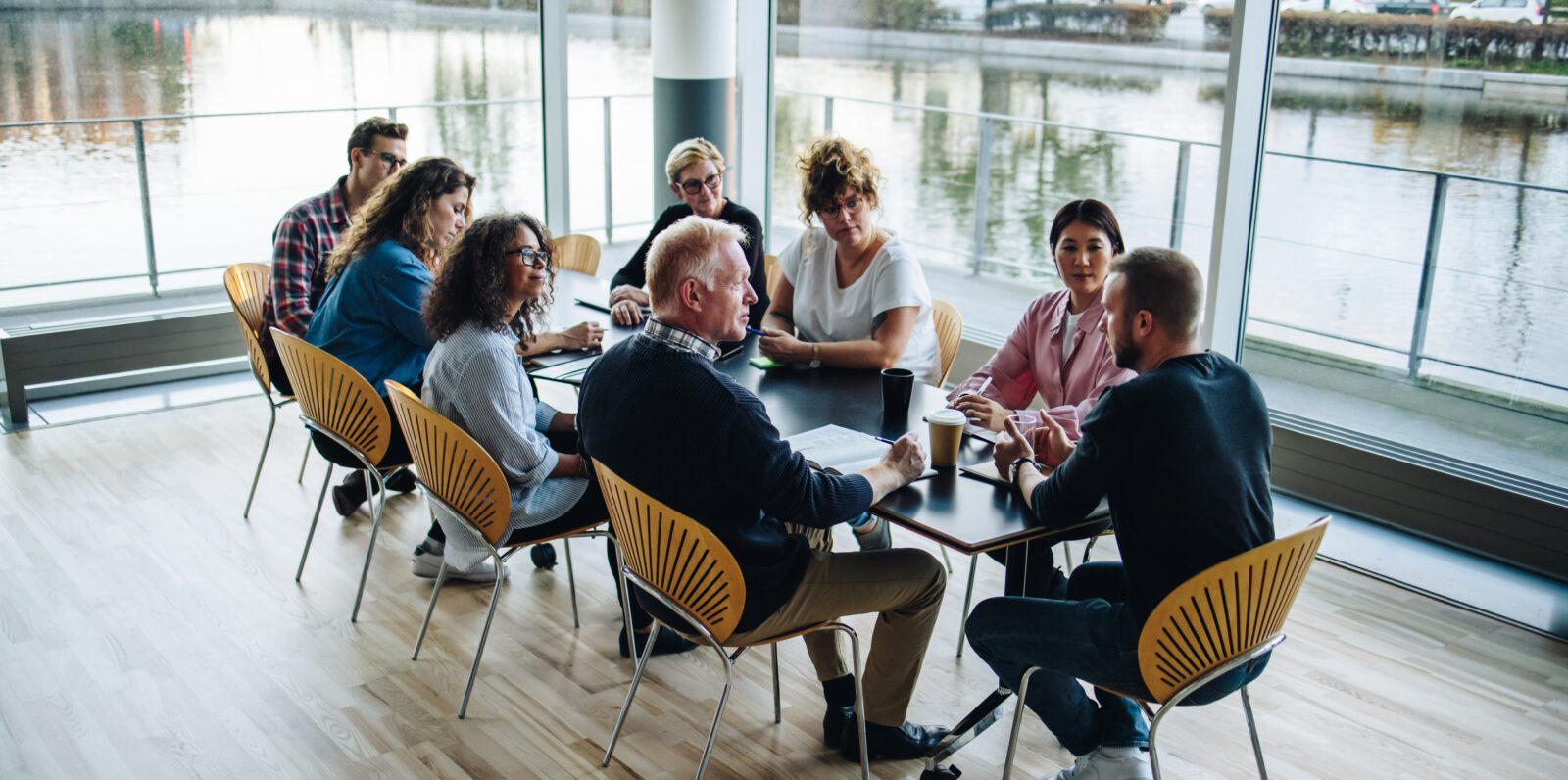 Eight people sit around a table in a modern meeting room with large windows overlooking water, discussing the advantages of peer groups for business leaders.