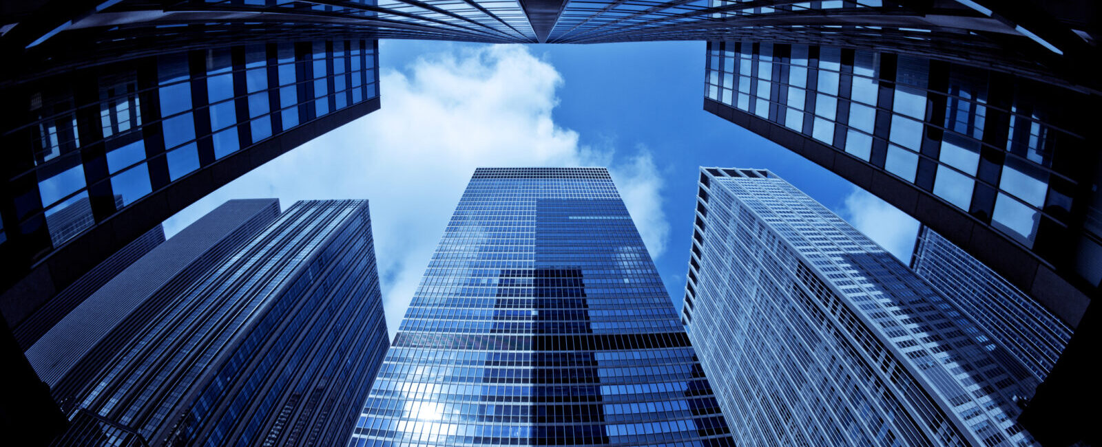 View from ground level looking up at several tall glass skyscrapers, reflecting the sky—much like pondering, What Happens to Your Bank-Owned Life Insurance?—all set against a backdrop of blue sky with drifting clouds.