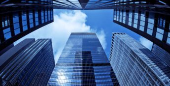 View from ground level looking up at several tall glass skyscrapers, reflecting the sky—much like pondering, What Happens to Your Bank-Owned Life Insurance?—all set against a backdrop of blue sky with drifting clouds.