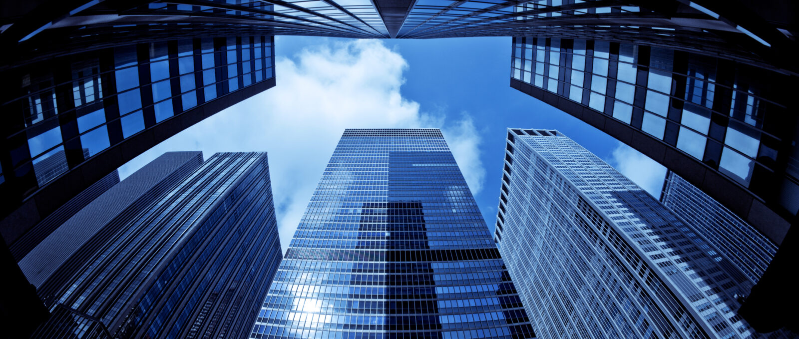 View from ground level looking up at several tall glass skyscrapers, reflecting the sky—much like pondering, What Happens to Your Bank-Owned Life Insurance?—all set against a backdrop of blue sky with drifting clouds.