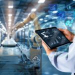 A person holds a tablet displaying data analytics and automation icons, overseeing robotic arms in a modern manufacturing facility—showcasing an essential AI business strategy for manufacturers in 2025.