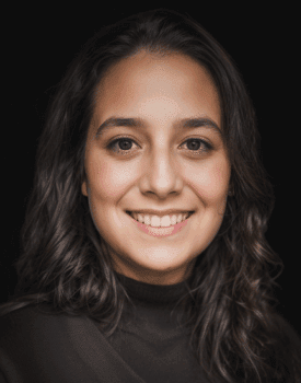 Carolina Vargas, with long, wavy brown hair and a dark top, smiles at the camera against a black background.