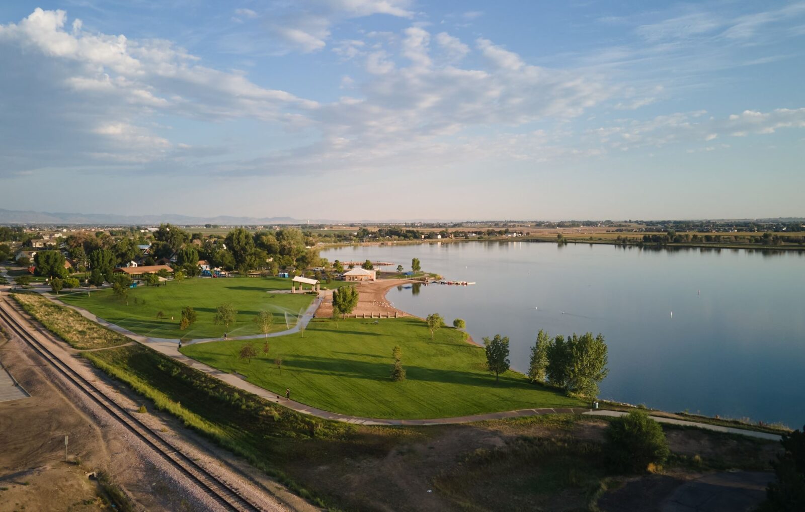 Aerial view of a lakeside park with green lawns, scattered trees, a sandy beach area, and adjacent railroad tracks under a partly cloudy sky.