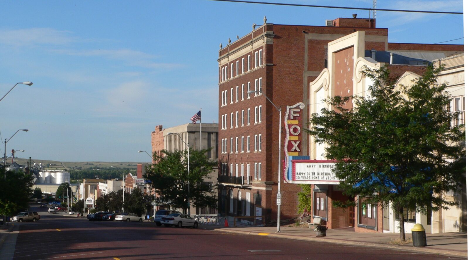 A small city street in McCook, NE with brick buildings, including the Fox theater with a marquee sign, trees lining the sidewalk, and a few cars parked along the curb.