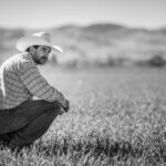A man in a cowboy hat and plaid shirt squats in a grassy field with mountains behind him, capturing the mental load of agriculture in black and white. For support, reach out to an ag hotline.
